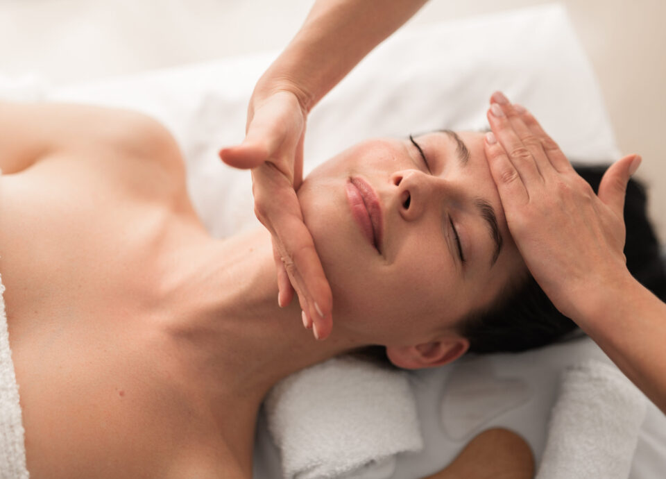 Relaxed young woman smiling while lying on table during face massage Top view of crop unrecognizable masseuse massaging face of content young female customer with dark hair and bare shoulders, lying on table and smiling with closed eyes during beauty procedure